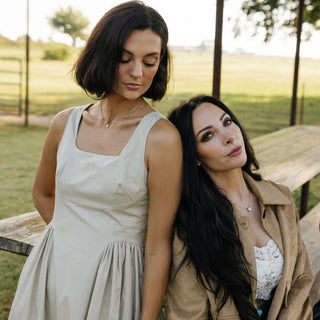 Two women standing close together outdoors with a wooden bench and greenery in the background.